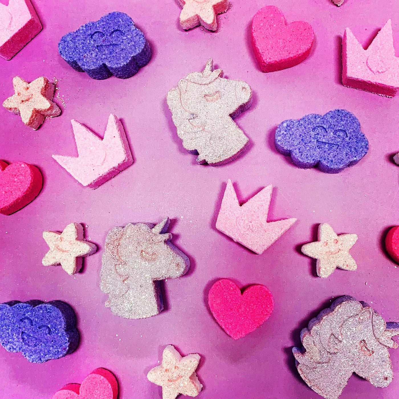 Colorful mini bath bombs shaped like unicorns, stars, and crowns on a pink background. Perfect for a magical bath experience.