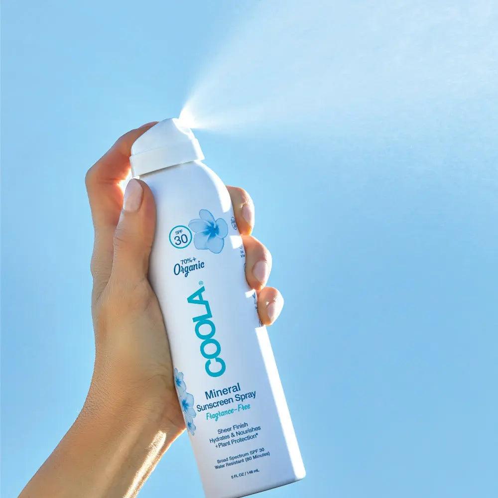 Hand holding COOLABio Mineral Body Sunscreen Spray SPF 30, fragrance-free and organic, spraying a fine mist against a blue sky.