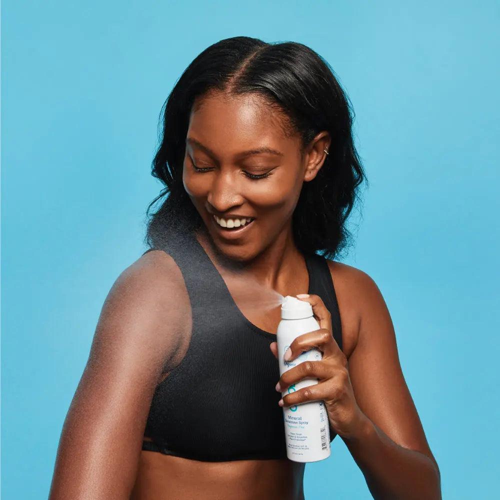 Woman applying Mineral Body Organic Sunscreen Spray SPF 30, enjoying its hydrating, fragrance-free formula against blue backdrop.