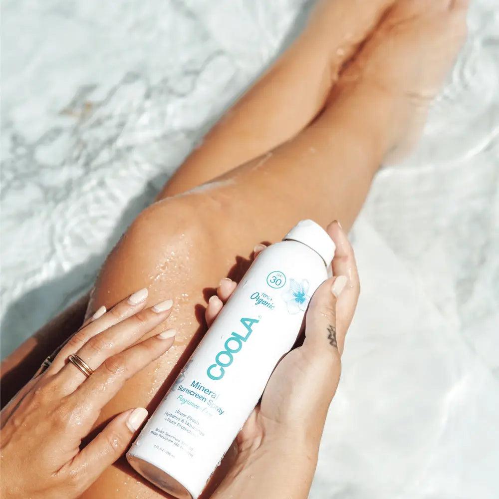 Woman applying mineral body organic sunscreen spray SPF 30 on leg by the water, showcasing fragrance-free sun protection.