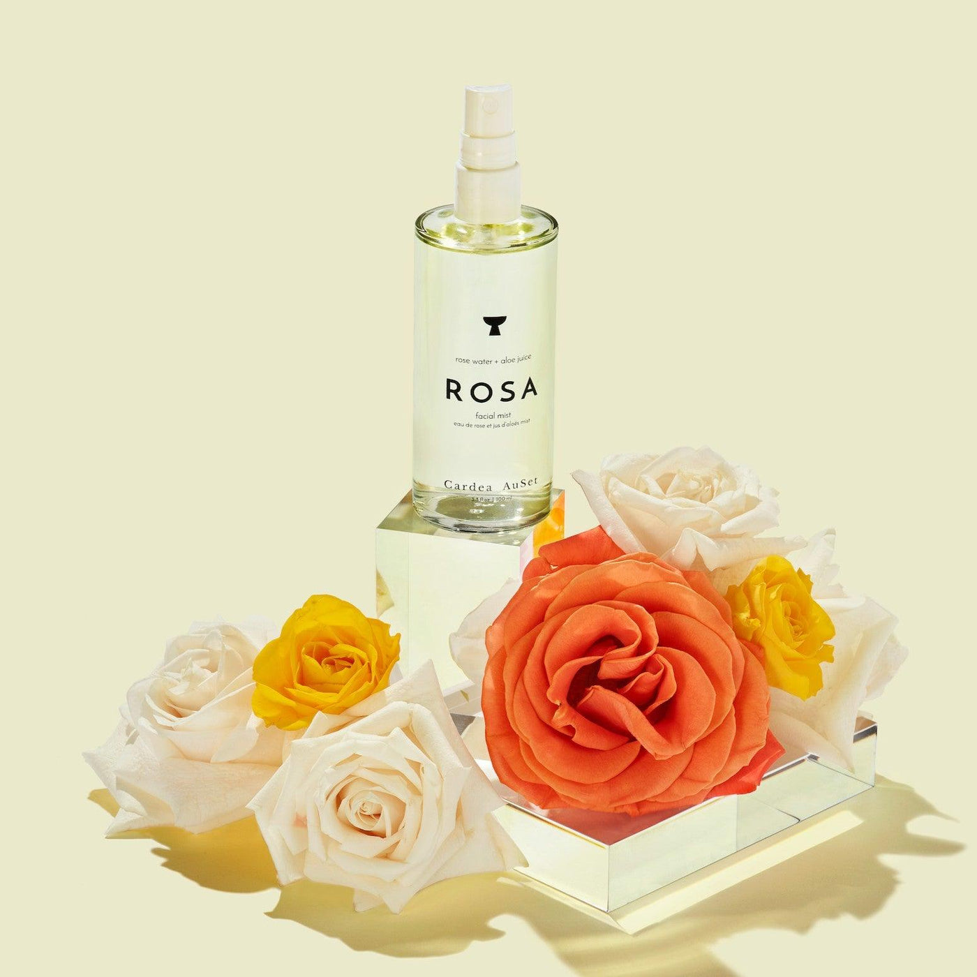 ROSA perfume bottle surrounded by white, yellow, and orange roses on a reflective surface