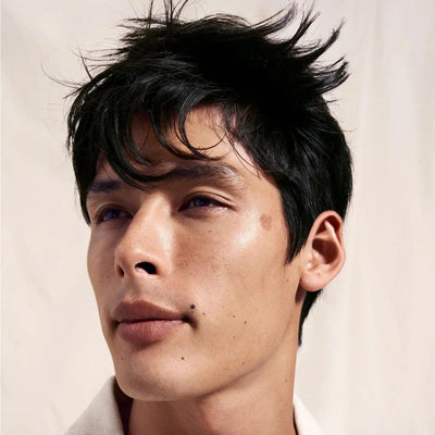 Young man with tousled black hair and a fresh, clean complexion posing against a light background.