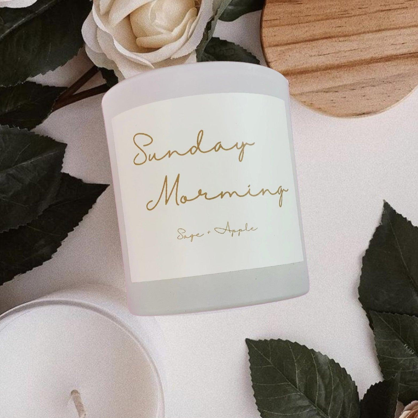 White glass candle jar labeled "Sunday Morning - Sage + Apple" with leafy backdrop and wooden lid on a table.