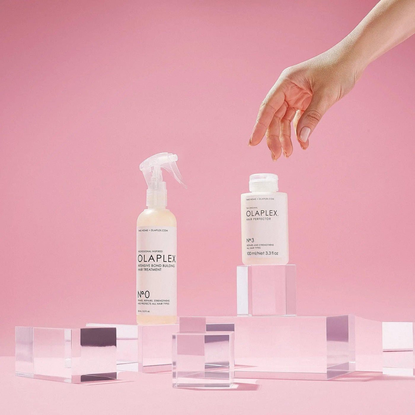 OLAPLEX No.0 and No.3 Repair Kit displayed on blocks with a hand reaching for them, against a pink background.